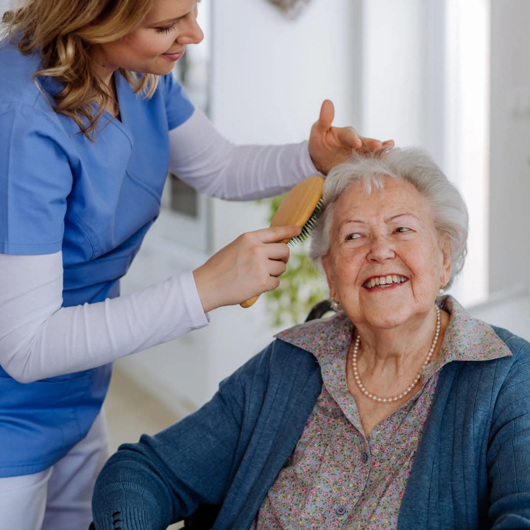 A caregiver brushing an older woman's hair as she smiles.