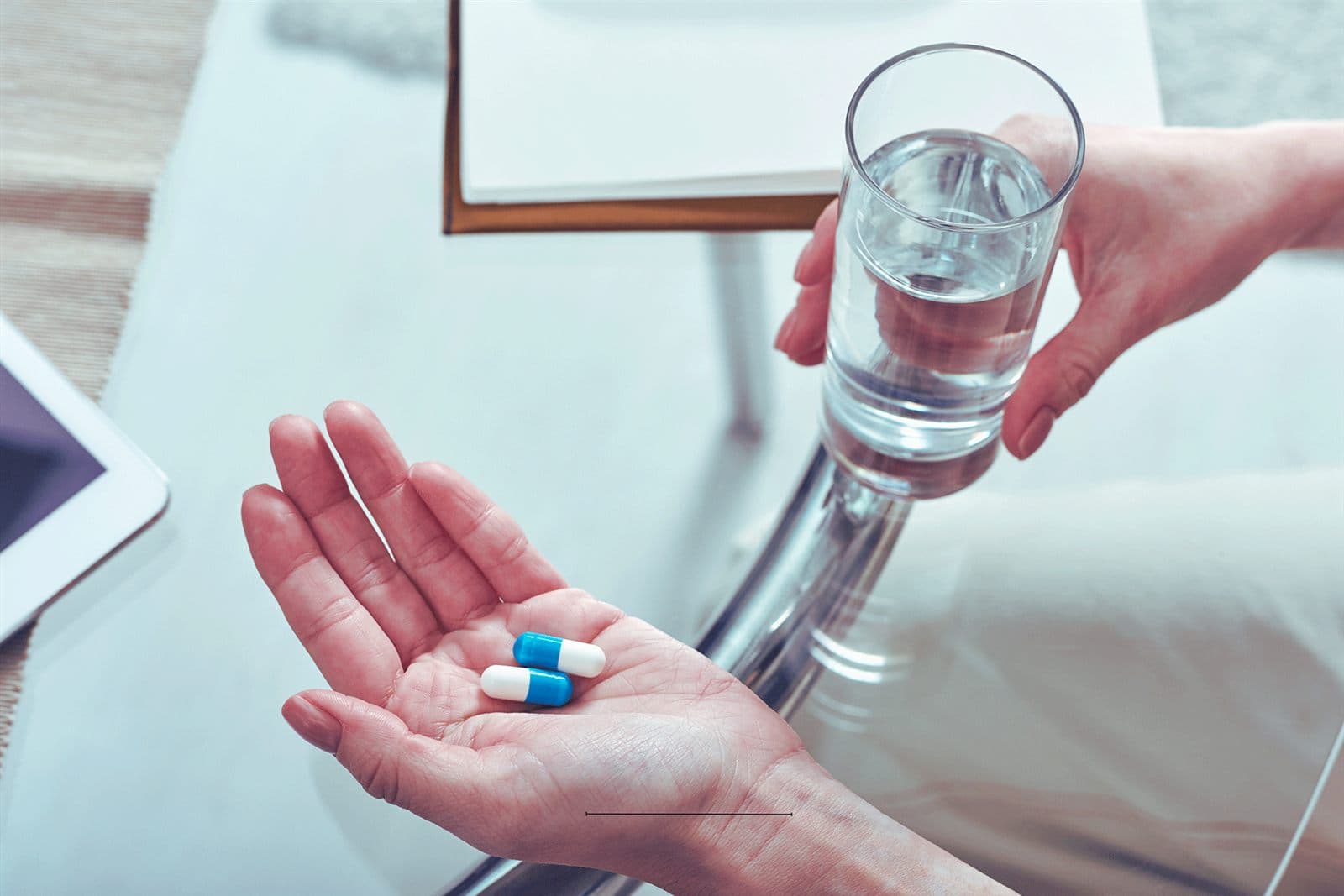 An open palm holding two capsules while another hand offers a glass of water.