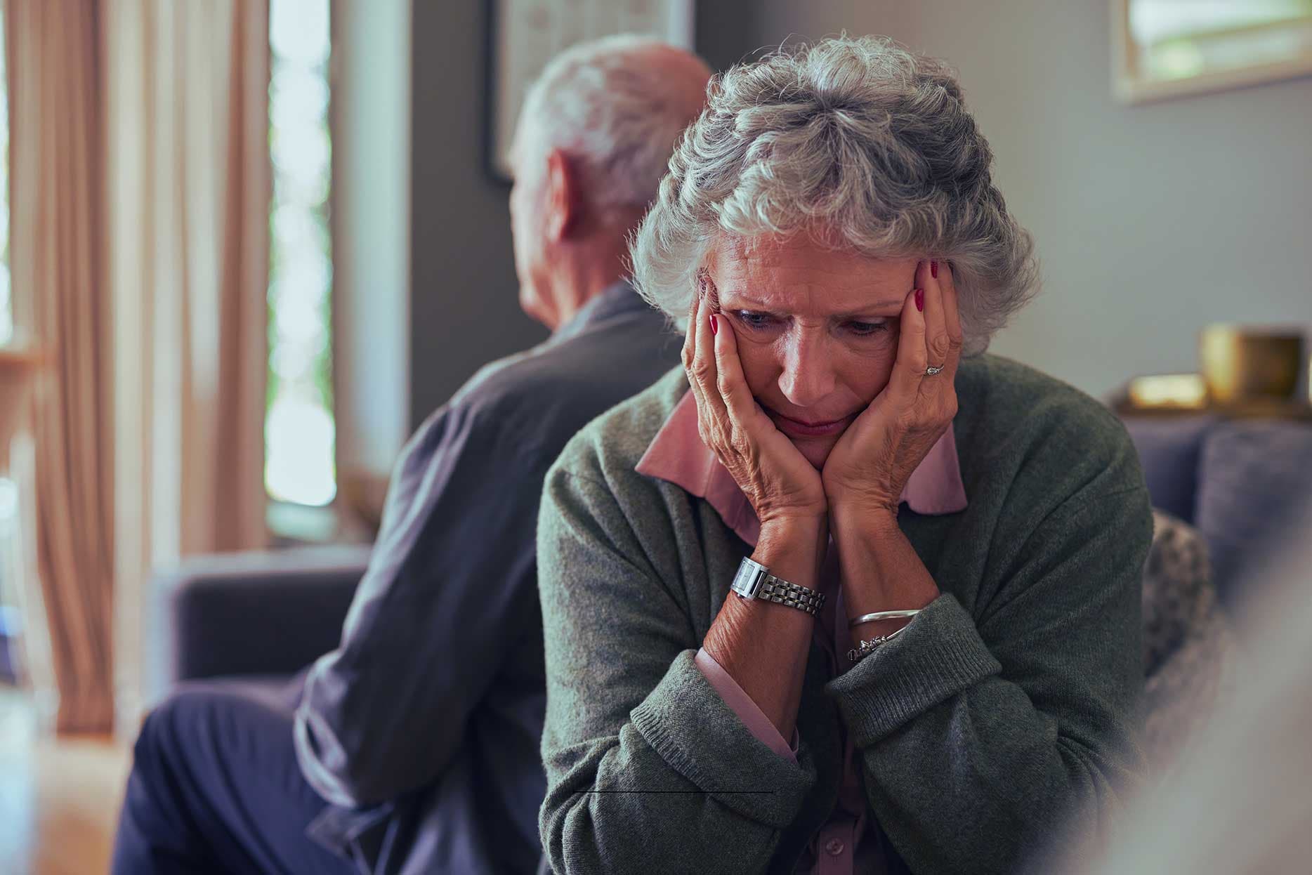 An older woman sitting with her hands to her face, looking troubled, an older man behind her.