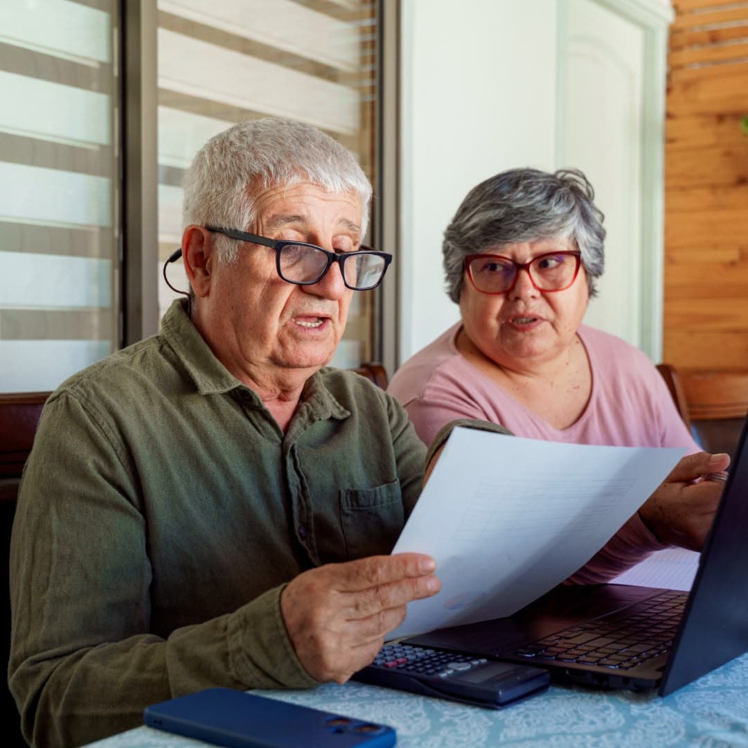An older couple reading a printed statement together beside a laptop at their dining table.