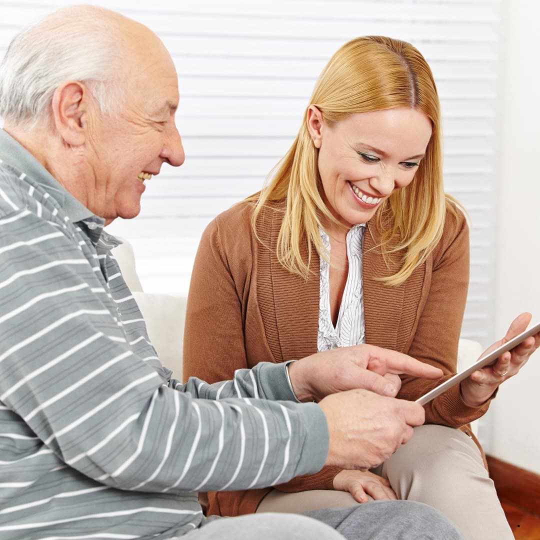 An older man and a woman sitting together, smiling as they look at a tablet he is holding.