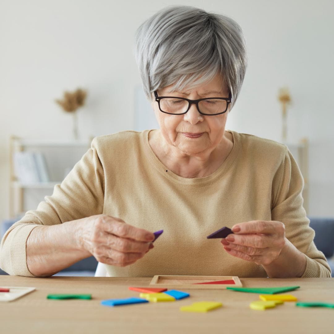 An older woman at a table working through a coloured shape puzzle, concentrating on two pieces in her hands.