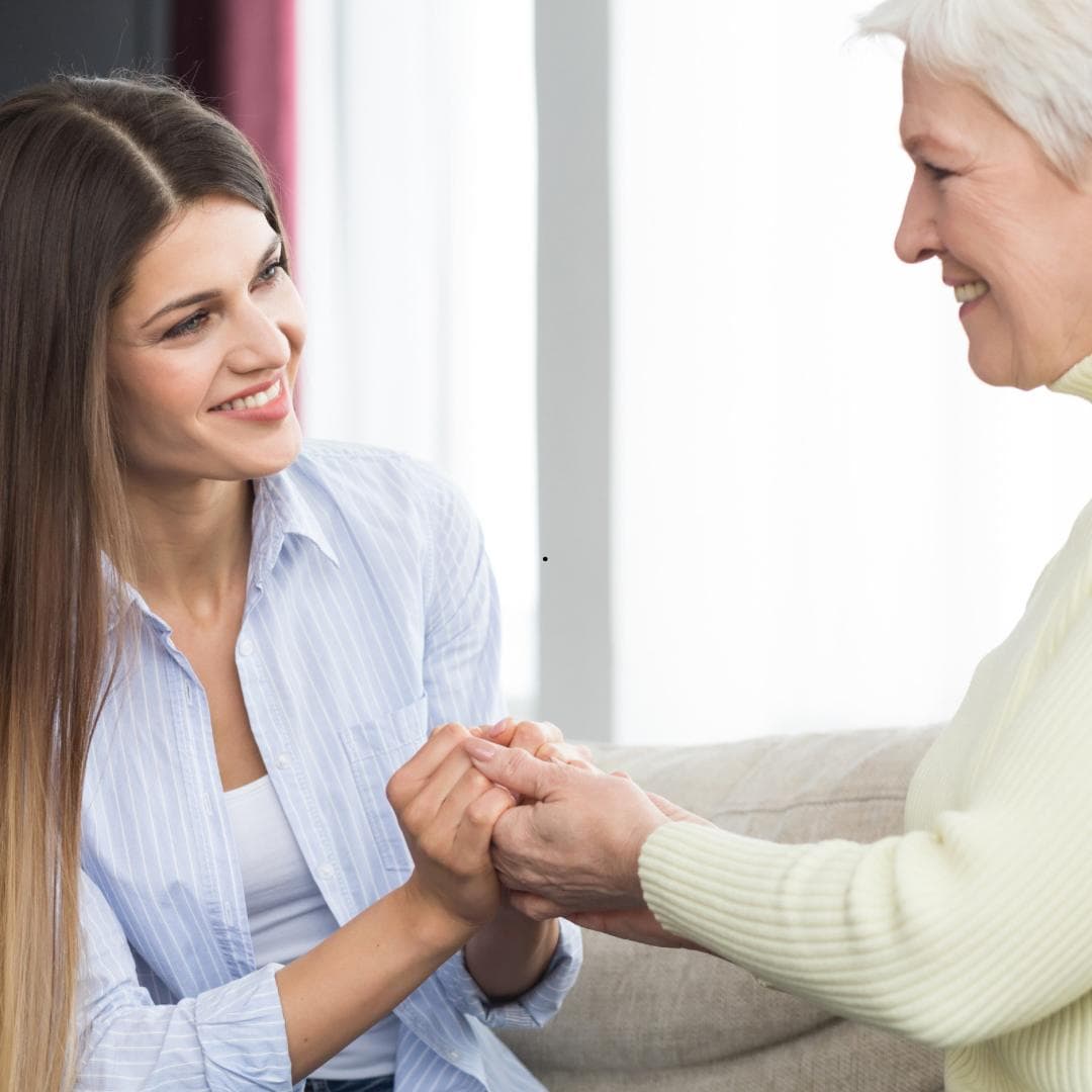 A young woman sitting on a sofa holding an older woman's hands, the two of them smiling at each other.