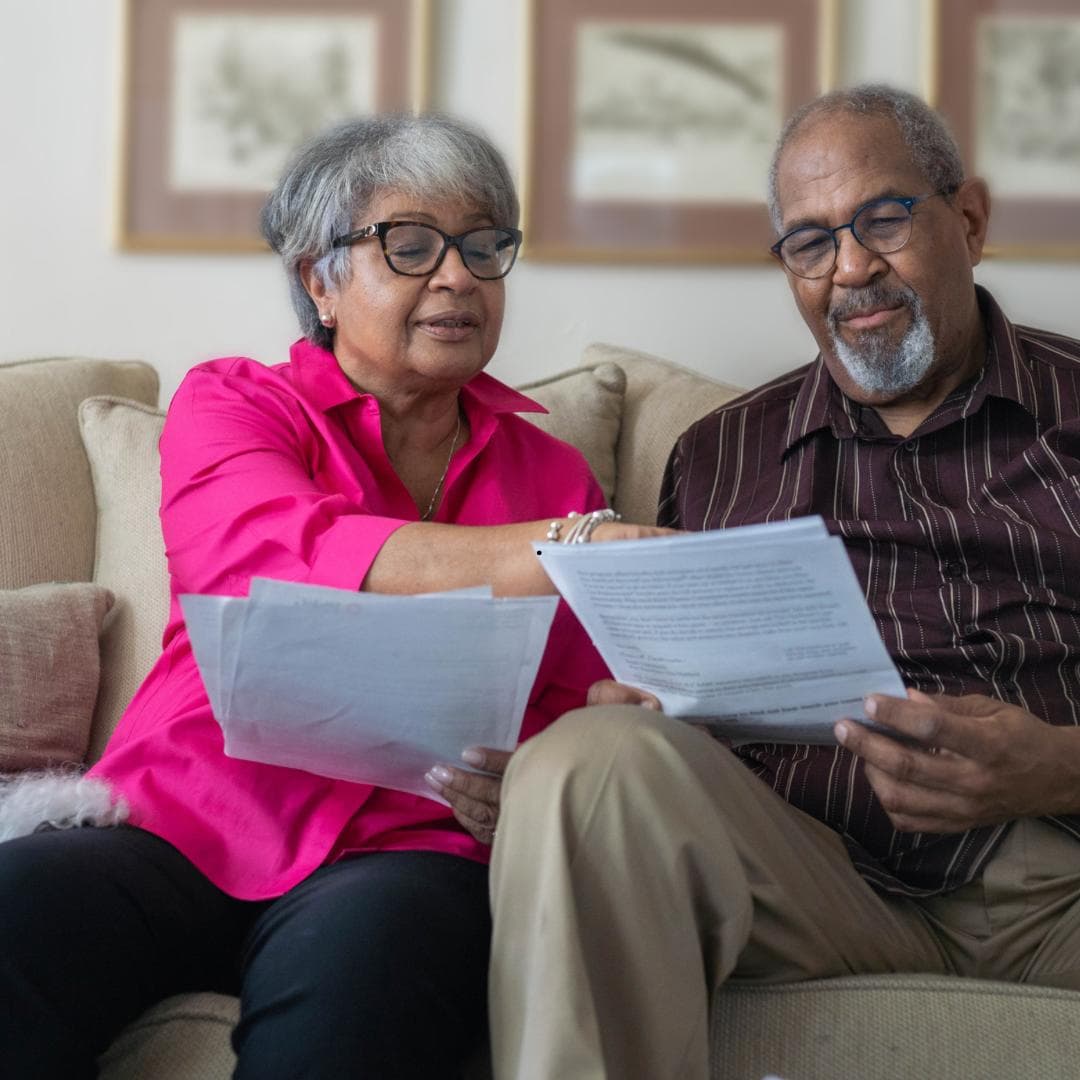 An older couple sitting on a sofa reading through printed documents together.