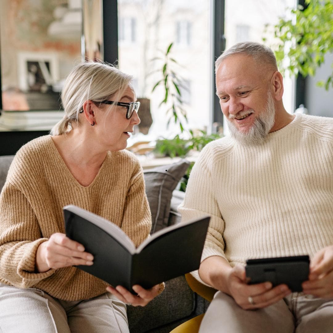 An older couple sitting together at home, one holding an open notebook and the other a tablet, talking something through.