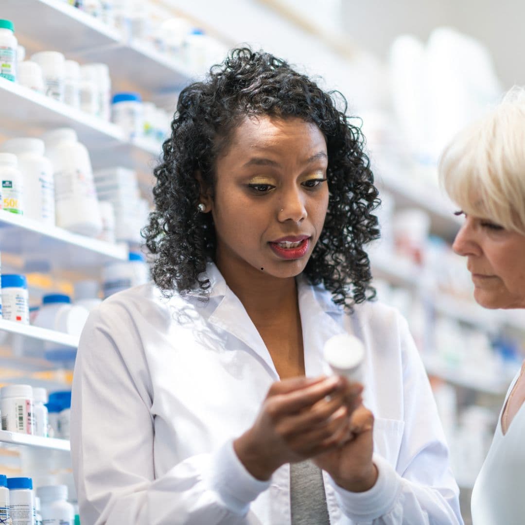 A pharmacist in a white coat reading the label on a pill bottle beside an older woman in a pharmacy aisle.