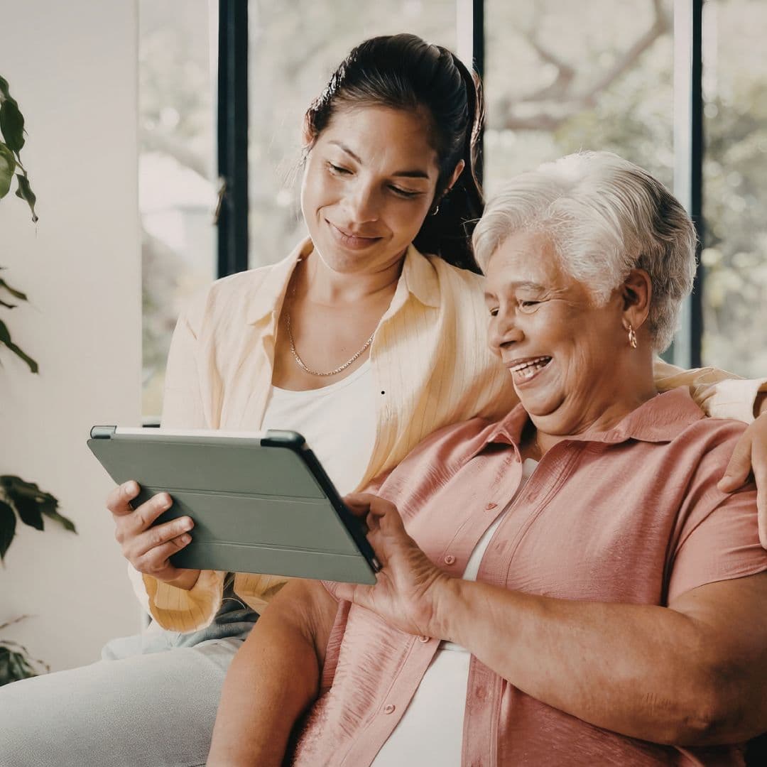A woman sitting with her arm around her mother, the two of them looking at a tablet together and smiling.