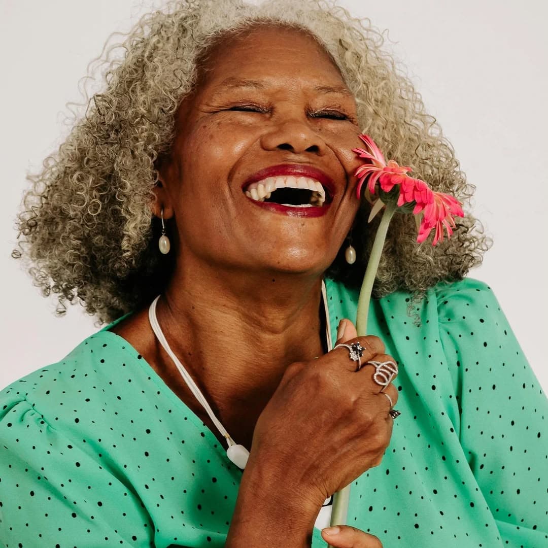 An older woman laughing with her eyes closed, holding a pink gerbera daisy up beside her face.