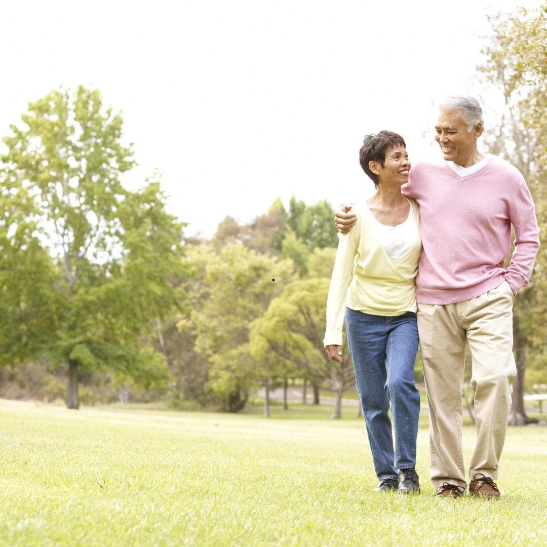 An older couple walking arm in arm across a wide lawn in a park, both laughing.