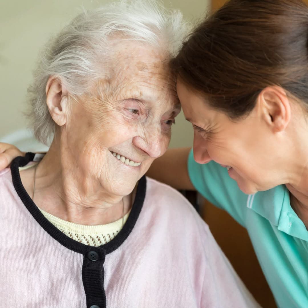 A caregiver resting her head against an older woman's forehead, both of them smiling.