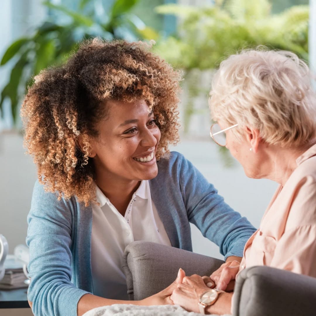 A woman leaning in to talk with an older woman, holding her hands, the two of them smiling at each other in a bright room full of plants.
