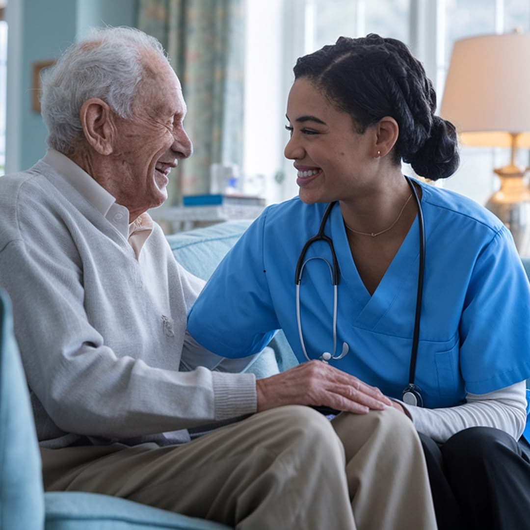 A nurse in scrubs sitting beside an older man in his living room, the two of them laughing together, her hand resting on his.